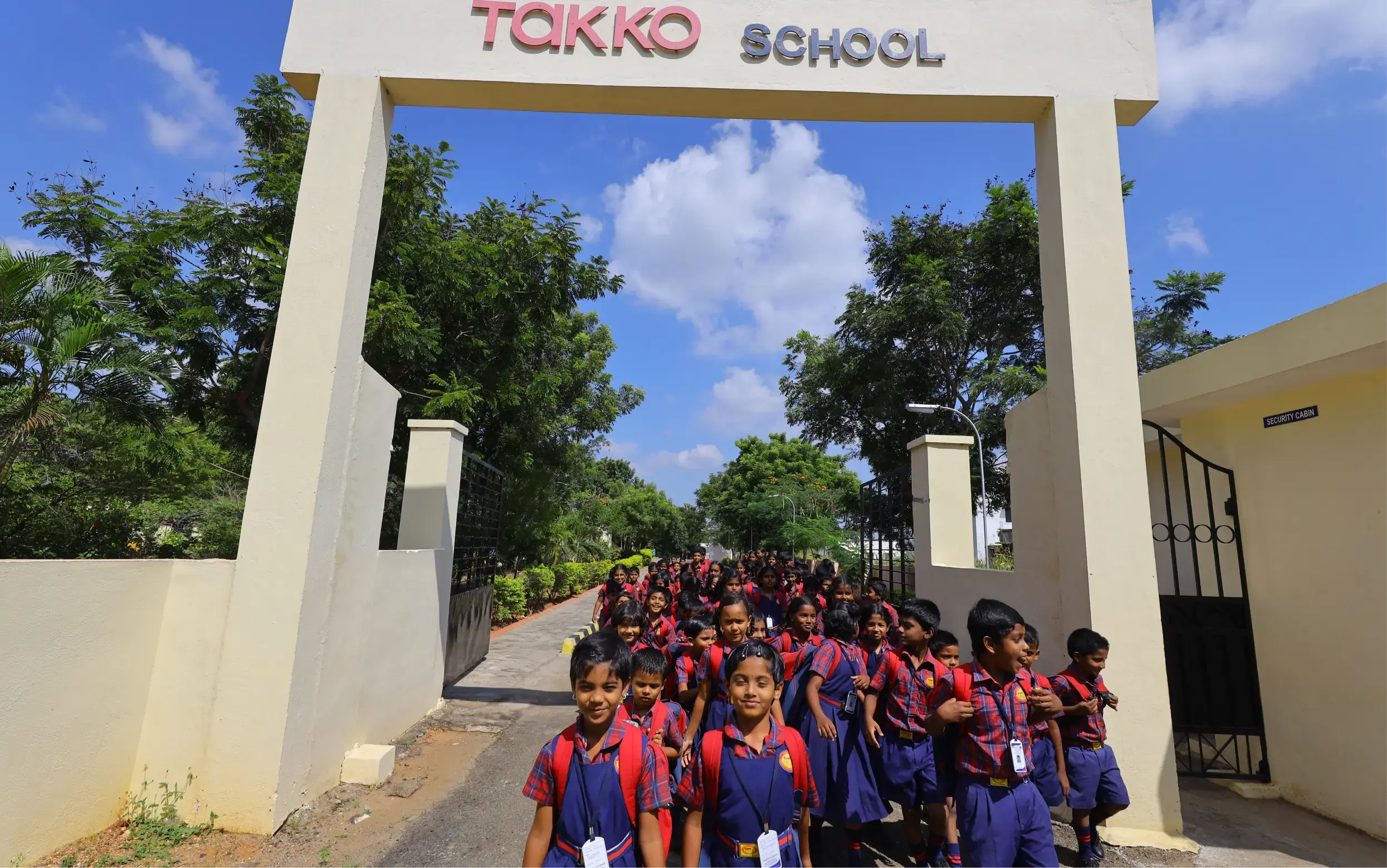 Eine große Gruppe junger Schüler in passenden rot-blauen Uniformen kommt an einem sonnigen Tag aus einem Schultor mit der Aufschrift „TAKKO SCHOOL“, im Hintergrund sind Bäume und ein blauer Himmel zu sehen.